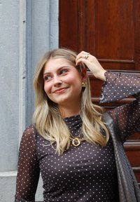 Blusa de tela negra transparente con puntos; mangas largas con puños acampanados. Joyería dorada, incluyendo collares en capas y pendientes de aro.