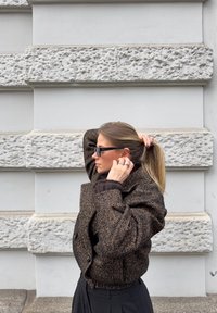 Γυναίκα με σκουρόχρωμο παλτό με σχέδια και γυαλιά ηλίου ρυθμίζει την ξανθή αλογοουρά της στο φόντο ενός υφής πετρόχτιστου τοίχου.