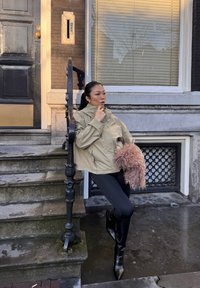 Femme en veste beige et bottes noires s'appuyant sur une rambarde près d'escaliers en pierre, tenant un sac rose duveteux devant un bâtiment.