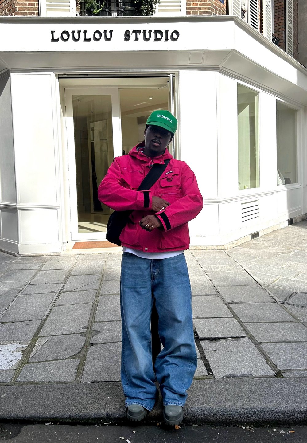Piros dzseki fekete részletekkel, fehér ing, laza kék farmernadrág és zöld sapka. Egy bolt előtt áll, amin a "LOULOU STUDIO" felirat látható.