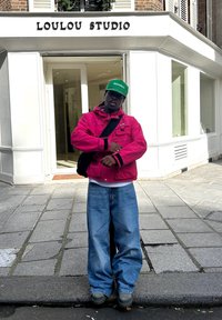 Piros dzseki fekete részletekkel, fehér ing, laza kék farmernadrág és zöld sapka. Egy bolt előtt áll, amin a "LOULOU STUDIO" felirat látható.