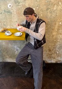 A young person wearing a grey waistcoat and trousers sits at a yellow table, holding a large white cup of coffee, with a croissant and a small cup beside them.