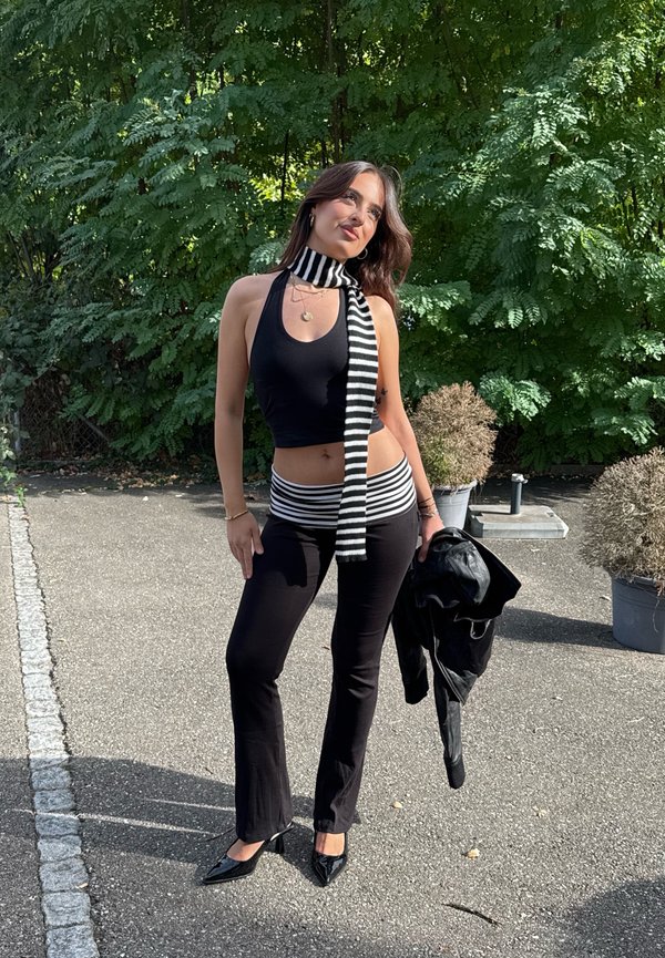 Mujer con top corto negro y pantalones con bufanda rayada y cinturilla, sosteniendo una chaqueta negra, de pie al aire libre sobre suelo pavimentado cerca de vegetación.