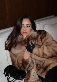 Cappotto di pelliccia marrone con una texture soffice, abbinato a guanti di pelle nera. Una piccola borsa nera poggia su un divano bianco.