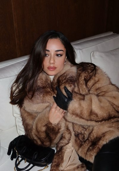 Abrigo de piel marrón con una textura suave, combinado con guantes de cuero negro. Un pequeño bolso negro descansa sobre un sofá blanco.