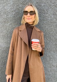 Ruskea villakangastakki, jossa on läpän muotoilu, yhdistettynä tumman väriseen poolopaitaan. Pitelee valkoista mukia, jossa on punainen suoja, ja käyttää tummia aurinkolaseja.