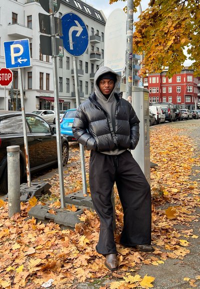 Chaqueta de pluma negra con capucha sobre un sudadera gris, combinada con pantalones holgados negros. De pie en una calle cubierta de hojas.