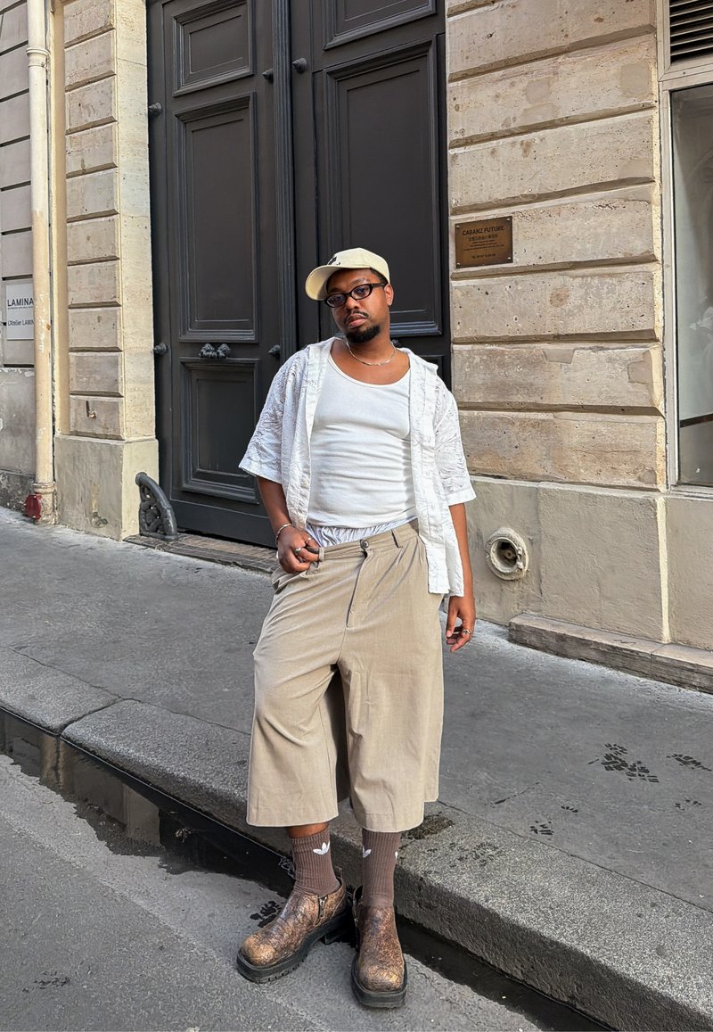 Homme portant une casquette beige, un débardeur blanc, une chemise à manches courtes ouverte, un pantalon large beige, des bottes à motifs et des chaussettes marron se tient dans une rue de la ville à côté d'une grande porte sombre.