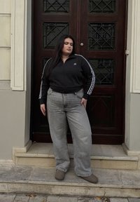 Μαύρο φερμουάρ μπουφάν με λευκές ρίγες, γκρι φαρδιά τζιν και γκρι υποδήματα, στέκεται σε πέτρινες σκάλες μπροστά από μια ξύλινη πόρτα.