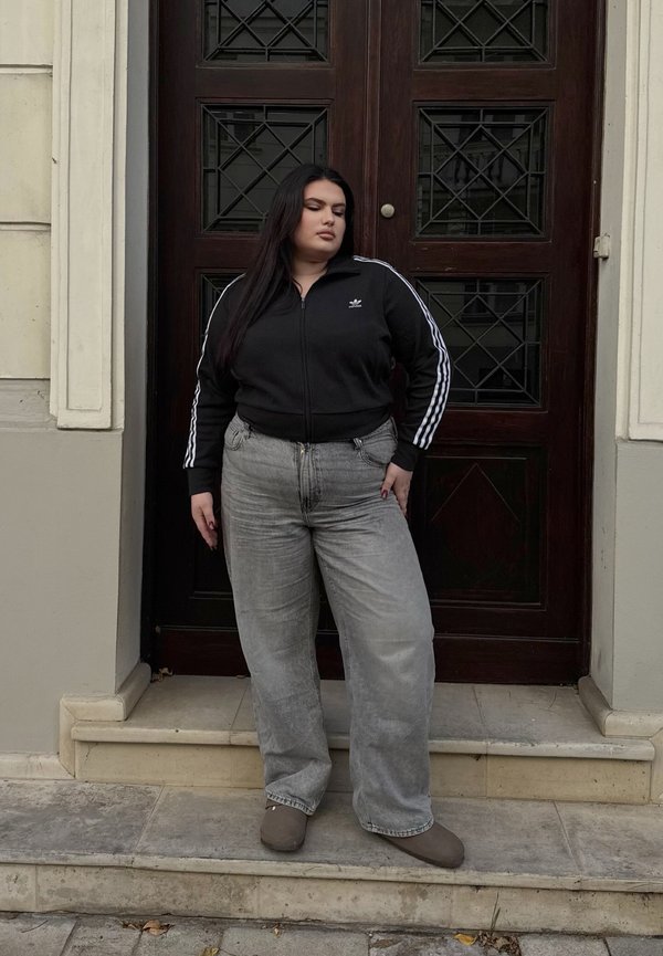 Veste noire zippée avec des rayures blanches, jeans baggy gris et chaussures grises, se tenant sur des marches en pierre devant une porte en bois.