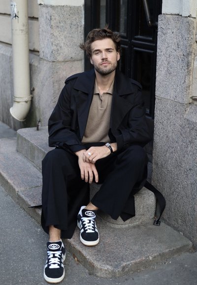 Abrigo negro sobre una camiseta beige, combinado con pantalones anchos negros y zapatillas negras con detalles en blanco. Sentado en unos escalones de piedra.