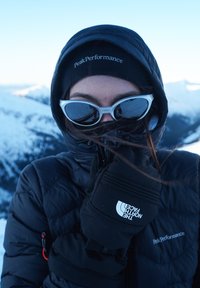 Person iført sort vinterjakke, handsker, hue og hvide solbriller, dækker ansigtet mod den kolde, sneklædte bjergbaggrund.