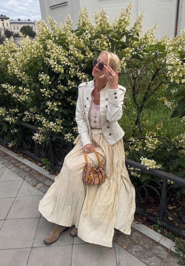 Femme en veste blanche et longue jupe beige assise près d'un buisson en fleurs, portant des lunettes de soleil et tenant un petit sac à main à motifs.