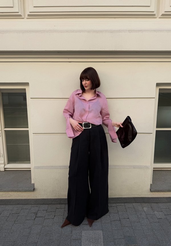 Vrouw met kort bruin haar die een roze gestreept overhemd draagt, ingestopt in een hoge wijde zwarte broek, houdt een zwarte clutch vast en staat bij een beige muur.