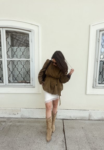Mujer con cabello largo y oscuro que lleva una chaqueta verde oliva, falda blanca con bordes de encaje y botas de ante beige, de pie entre dos ventanas con rejas decorativas.