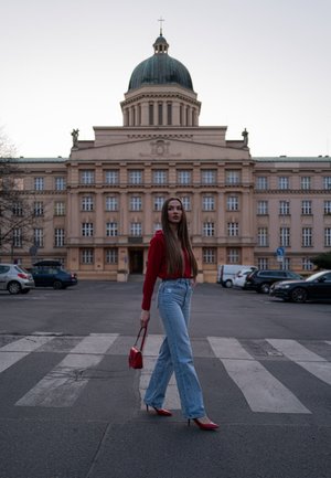 Sudadera roja, jeans acampanados de color azul claro, tacones altos rojos y un pequeño bolso rojo, caminando por un paso de peatones con un edificio histórico al fondo.
