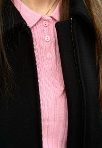Femme portant un polo en maille rose à trois boutons sous une veste noire partiellement zippée, avec de longs cheveux raides visibles.
