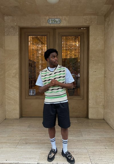 Chaleco de punto con patrones de olas verdes y negros sobre una camiseta azul claro, combinado con pantalones cortos negros, calcetines blancos y mocasines negros.