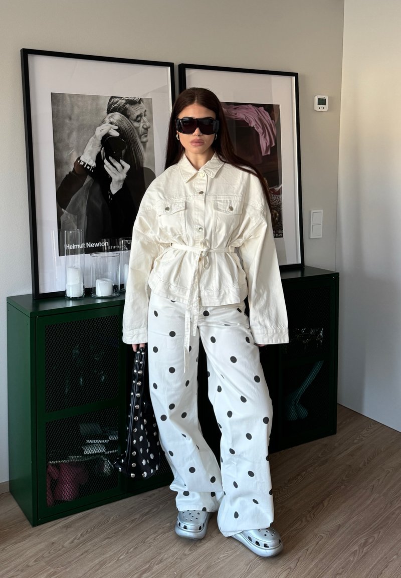 Γυναίκα με oversized λευκό σακάκι, λευκό παντελόνι με πουά, μεγάλα γυαλιά ηλίου, κρατά τσάντα με καρφιά, στέκεται σε εσωτερικό χώρο δίπλα σε καδραρισμένες ασπρόμαυρες φωτογραφίες.