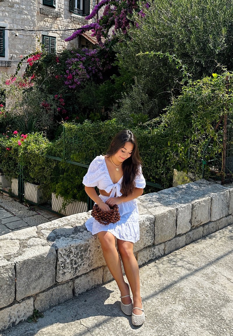 Λευκό κεντημένο τοπ με κοντό μήκος και ταιριαστή φούστα με ριγωτό τελείωμα. Κρατάει μια διακοσμητική ομάδα καφέ μούρων, καθισμένη σε έναν τοίχο από πέτρες.