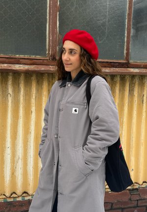 Mujer con boina roja y abrigo gris con las manos en los bolsillos, de pie frente a una pared amarilla corrugada y una ventana antigua.