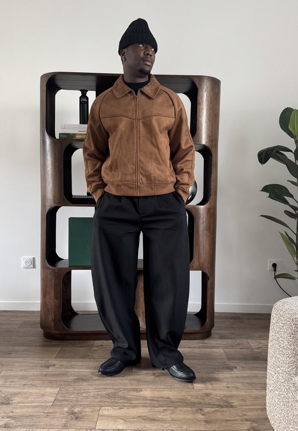 Hombre con chaqueta marrón, pantalones negros, gorro negro y zapatos, de pie con las manos en los bolsillos en una habitación moderna con estanterías de madera y una planta.