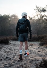 Sac à dos noir, chemise noire à manches longues, short gris, chaussettes blanches avec des rayures noires, chaussures orange et blanches, marchant sur un sentier sablonneux en pleine nature.
