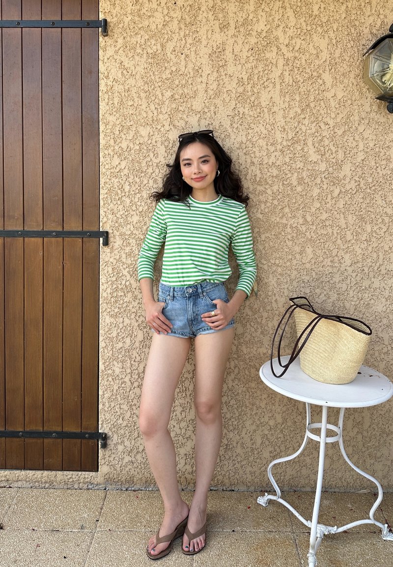 Jeune femme en haut rayé vert et blanc et short en jean se tient contre un mur beige texturé près d'une porte en bois et d'une petite table ronde avec un sac en paille.