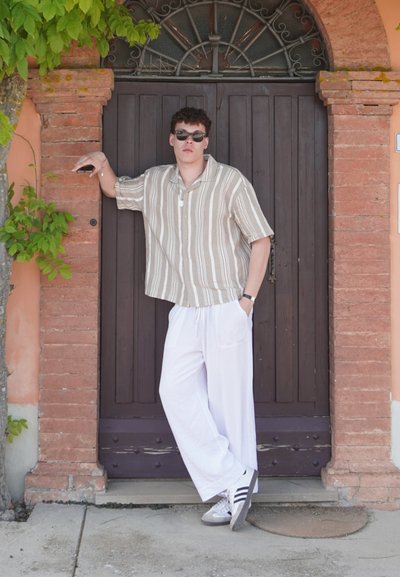 Camisa de manga corta a rayas en beis y blanco, combinada con pantalones blancos holgados. Zapatillas grises con rayas negras. De pie contra una puerta de madera.