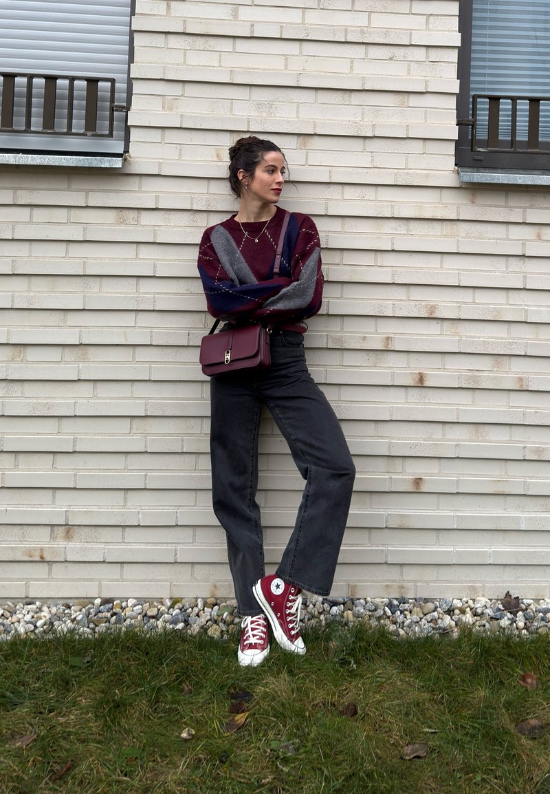 Nuori nainen, jolla on tummat hiukset nutturalla, pukeutuneena väriblokkineuleeseen, mustiin farkkuihin, punaisiin lenkkareihin ja viininpunaiseen olkalaukkuun, seisoo valkoista tiiliseinää vasten.