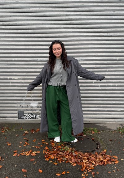 Abrigo largo gris, suéter corto gris con botones, pantalones verdes de pierna ancha, zapatillas blancas. Bolsa transparente con patrones circulares. Hojas caídas en el suelo.