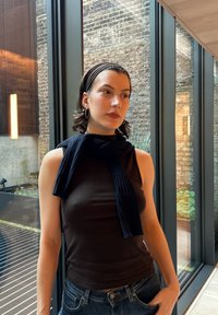 Écharpe noire côtelée enroulée autour du cou, associée à un haut sombre sans manches. Cheveux courts et grandes boucles d'oreilles argentées visibles.