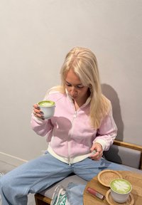 Jeune femme aux cheveux blonds, vêtue d'une veste rose et de jeans, assise en tailleur, tenant une tasse de latte au matcha avec un art de latte sur une table en bois.