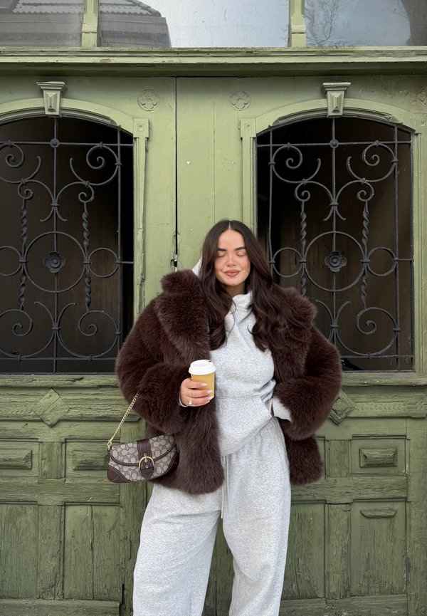 Femme en manteau de fourrure marron et pantalon de survêtement gris tenant une tasse de café, debout les yeux fermés devant une porte en bois verte avec des grilles en fer noir.