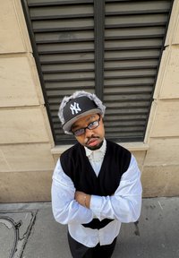 Homme portant une casquette noire des New York Yankees avec bordure en fourrure, un gilet noir, une chemise blanche, des lunettes et un collier de perles, debout contre un mur en pierre.