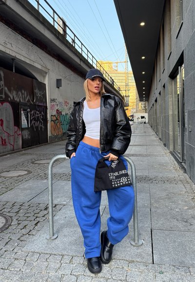Chaqueta negra acolchada, top corto blanco, pantalones de chándal azules, gorra negra, sosteniendo una bolsa de tela negra con texto blanco. Entorno urbano con graffiti.