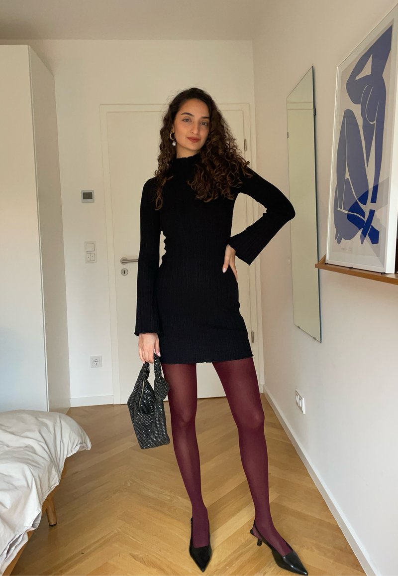 Schwarzes, geripptes Strickkleid mit langen, ausladenden Ärmeln, kombiniert mit burgunderfarbenen Strumpfhosen und spitzen schwarzen High Heels. Hält eine strukturierte schwarze Handtasche.
