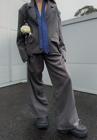 Personne portant un costume gris oversize, une cravate bleue à motifs, des sabots noirs à plateforme, et un porte-clés avec une peluche attachée à la veste.