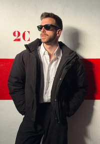Homme portant des lunettes de soleil noires, une veste matelassée noire, une chemise blanche et un pantalon noir, debout contre un mur blanc avec une bande rouge et le texte "2C".