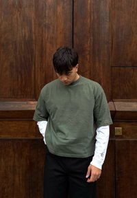 Jeune homme aux cheveux noirs regardant vers le bas, portant une chemise verte à manches courtes par-dessus une chemise blanche à manches longues, debout devant une porte en bois.