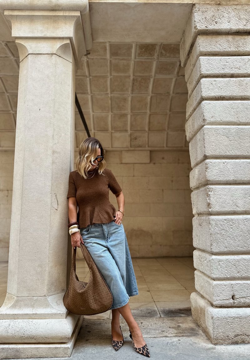 Donna con top marrone, gonna di jeans e tacchi a stampa leopardata appoggiata a una colonna di pietra, con una borsa intrecciata marrone in mano, sotto un soffitto ad arco in pietra.