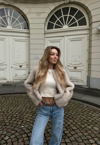 Casaco de pelo sintético bege sobre uma blusa branca de algodão cortada, combinado com calças de ganga largas de tom azul claro e um cinto castanho, de pé sobre calçada.