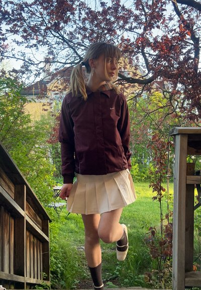 Chaqueta burdeos con capucha y logo, falda plisada color crema, calcetines negros y zapatos blancos. Entorno natural con hierba verde y árboles.