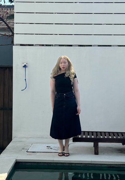 Top negro off-the-shoulder combinado con una larga falda de mezclilla negra, de pie junto a una piscina. El fondo presenta una pared de color claro y paneles de madera.