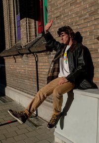 Svart, blank ytterjakke med brun krage over en hvit T-skjorte med et fargerikt grafisk design. Beige bukser og brune, snørebåndsboots med gule snøringer.