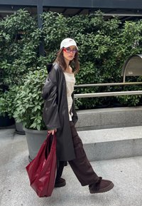 Femme portant une casquette blanche, des lunettes de soleil rouges, un manteau en cuir noir et un pantalon marron, marchant en extérieur avec un grand sac rouge et des plantes en arrière-plan.