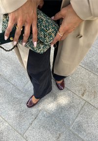 Τσάντα πλέξης σε περισσό χρώμα με χρυσά αξεσουάρ, μπεζ εξωτερικό παλτό, σκούρα τζιν και μπορντό παπούτσια σε ματ φινίρισμα. Χέρια κοσμημένα με δαχτυλίδια και καλοσχηματισμένα νύχια.