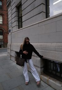 Zwarte oversized trui, witte wijd uitlopende broek, bruine handtas met patroon en beige plateauzolen. Staand tegen een stenen muur.