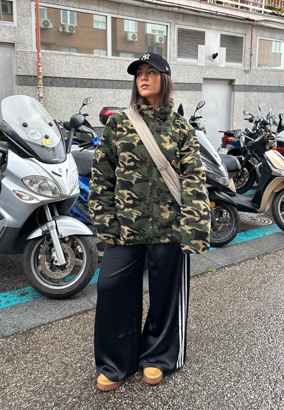 Chaqueta de forro polar de camuflaje con cuello alto, pantalones de pierna ancha negros y botas de plataforma beige, complementada con una gorra negra.