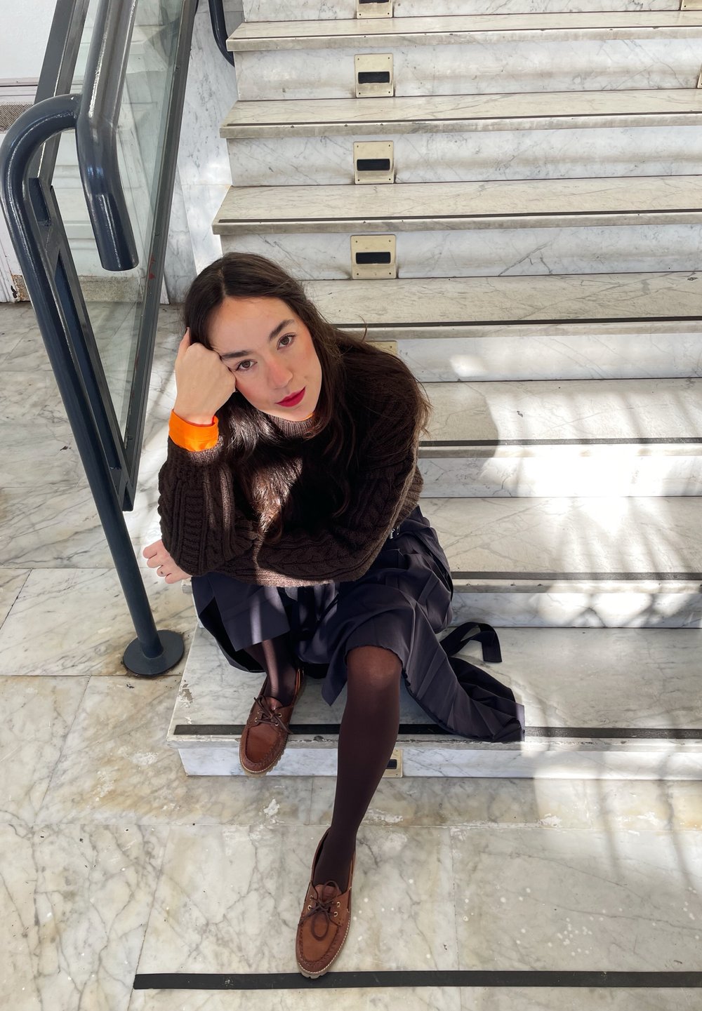 Persona seduta su scale di marmo, indossando un maglione di lana marrone, una gonna blu navy e scarpe marroni. Gli accessori includono un braccialetto arancione.